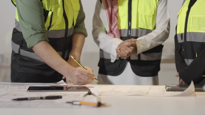 تقديم اعضاء الفريق للملاحظات قبل بداية التنفيذ , رجل اجنبي يرتدي الزي الكاجوال يقوم برسم المخطط الخاص بالعمل , مدير الفريق يقدم التعديلات , امرأة عربية خليجية سعودية ترتدي عباءة سوداء تشير الى شئ ما , الوقوف حول المكتب , سماع النصائح و الارشادات