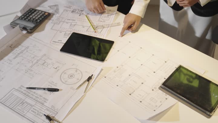 شرح تفصيلي للتخطيط المعماري بين اعضاء الفريق , مهندس عربي خليجي سعودي يرتدي الثوب الابيض يقوم بتقديم المعلومات الخاصة بالرسومات الهندسية , مهندسة عربية خليجية سعودية ترتدي العباءة السوداء تشير باصبع السبابة الى الاوراق على الطاولة , الادوات الهندسية المستخدمة في الرسم