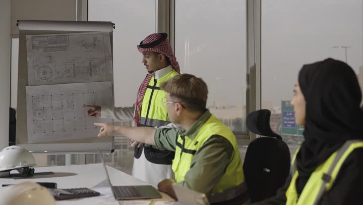 مهندس اجنبي يرتدي الزي الكاجوال يشير باصبع السبابة الى جزء ما في المخطط الهندسي , رجل عربي خليجي سعودي يرتدي الثوب الابيض و السترة الواقية العاكسة للضوء يقوم بشرح الملاحظات الخاصة بالرسومات المعمارية , امرأة عربية خليجية سعودية محجبة ترتدي العباءة السوداء تستمع بانصات الى الشرح التوضيحي , مفهوم العمل الجماعي