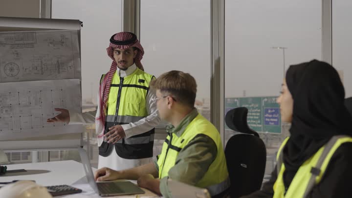 مهندسة عربية خليجية سعودية محجبة ترتدي العباءة السوداء تشير باصبع السبابة الى شئ ما في المخطط الهندسي , مهندس عربي خليجي سعودي يرتدي الثوب الابيض  والشماغ الملون يقوم بتوضيح المخططات الهندسية لباقي فريق العمل , رجل اجنبي يرتدي الزي الكاجوال يقوم بتسجيل الملاحظات على الحاسوب المتنقل