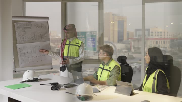 الاستماع بانصات لتوجيهات مدير الفريق , مهندس عربي خليجي سعودي يرتدي الثوب الابيض و الشماغ الملون يقف امام الرسومات الهندسية المعلقة للشرح والتوضيح , امرأة عربية خليجية سعودية محجبة ترتدي عباءة سوداء تجلس امام الجهاز اللوحي , رجل اجنبي يرتدي الزي الكاجوال يشير الى جزء ما في الرسم الهندسي