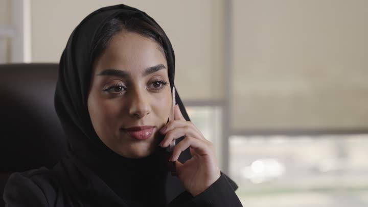 اجراء المكالمات الهاتفية الخاصة بالعمل من المكتب الهندسي , امرأة عربية خليجية سعودية محجبة ترتدي العباءة السوداء تقدم الاستشارات الهندسية باستخدام الهاتف المحمول , ايماءات السعادة و الابتسامة