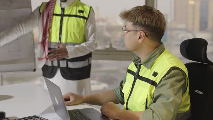 مناقشة عملية بين الزملاء داخل الفريق , مهندس عربي خليجي سعودي يرتدي الثوب الابيض و الشماغ الملون يوضح المخططات الهندسية , رجل اجنبي يرتدي الزي الكاجوال و السترة الواقية العكسة للضوء يتحدث مع باقي اعضاء فرق العمل , مفهوم العمل الجماعي , الحاسوب المتنقل على الطاولة