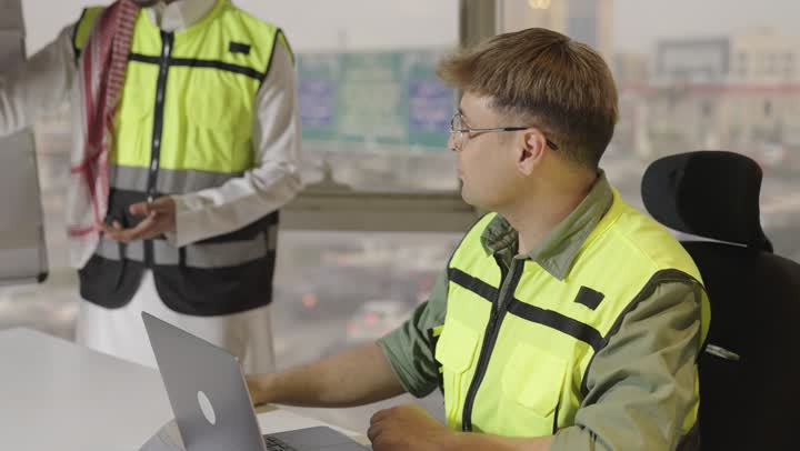 مهندس اجنبي يرتدي الزي الكاجوال يحرك رأسه بالموافقة على كلام المهندس المعماري , مهندس عربي خليجي سعودي يرتدي السترة الواقية العاكسة للضوء و الثوب الابيض يقوم بشرح المخططات المعمارية لباقي زملاء العمل , امرأة عربية خليجية سعودية تشير باصبع السبابة الى الرسم الهندسي المعلق , الحاسوب المتنقل على الطاولة