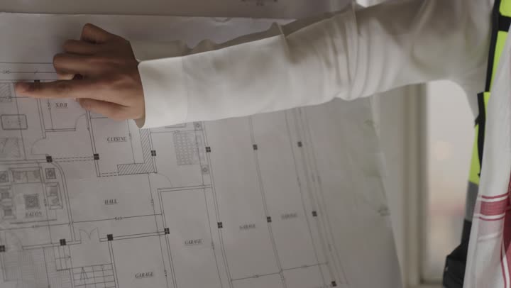 توضيح و شرح التخطيط الهندسي لباقي زملاء العمل , مهندس عربي خليجي سعودي يرتدي الثوب الابيض و الشماغ الملون يشير باصبع السبابة للرسومات الهندسية المعلقة على طاولة العرض , تصوير مقرب , ارتداء السترة الواقية العاكسة للضوء