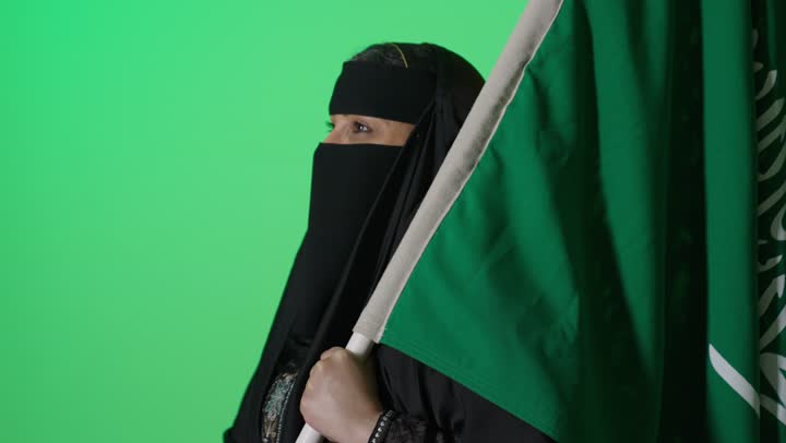 Pride in the achievements of the nation, the slogan of national identity and the banner of Islam and monotheism, a portrait of an elderly Saudi Arabian Gulf woman in a niqab wearing a black abaya holding the founding flag in her hand, looking with gestures of pride, commemorating the establishment of the first Saudi state on February 22, with a green background.