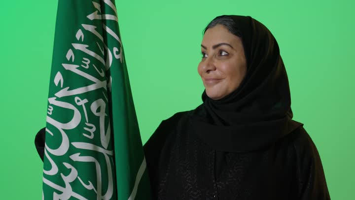 شعار الهوية الوطنية وراية الإسلام والتوحيد، بورتريه لامراه عربيه خليجيه سعوديه كبيرة بالسن ترتدي العباءة السوداء تحمل بيدها علم التأسيس, تنظر بايماءات الفخر، ذكرى تأسيس الدولة السعودية الأولى 22 فبراير، الاعتزاز بإنجازات الوطن، كروما، خلفية خضراء