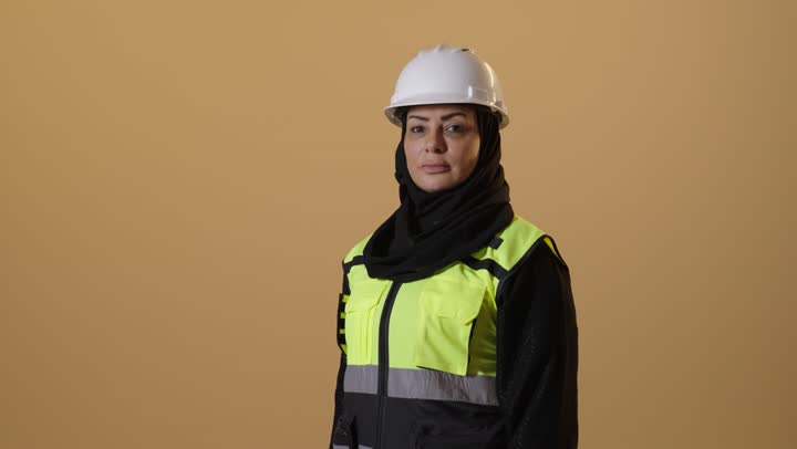 The concept of engineering project management, women's jobs and professions, working in the engineering sector, standing with gestures of confidence, a portrait of an elderly Arab Gulf Saudi female architect wearing a safety helmet and vest, standing with arms crossed, beige background.