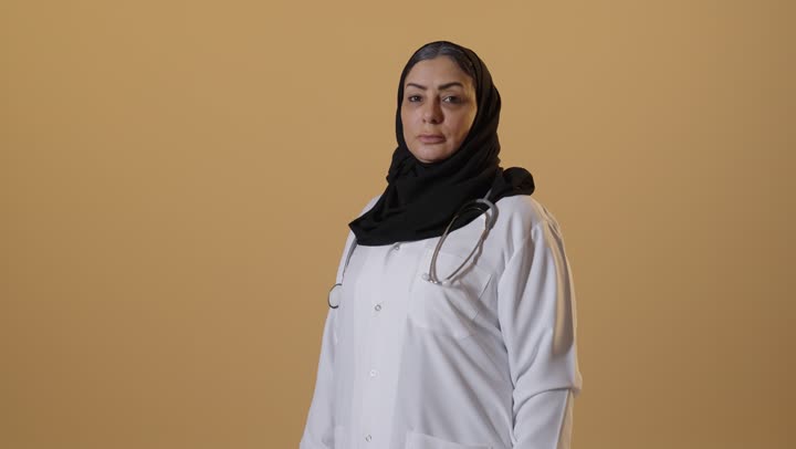 تقديم الاستشارات الطبية والخدمات للمرضى، عمل النساء في مجال الرعاية الصحية، بورتريه لامرأة عربية خليجية سعودية كبيرة بالسن محجبة ترتدي المعطف الطبي وسماعة الفحص، خلفية بيج