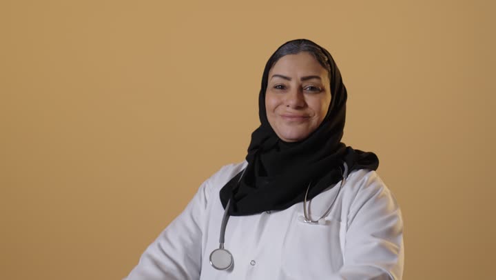 Women's work in the healthcare field, providing medical consultations and services to patients, a portrait of an elderly Saudi Gulf Arab woman wearing a hijab, dressed in a medical coat and stethoscope, beige background.