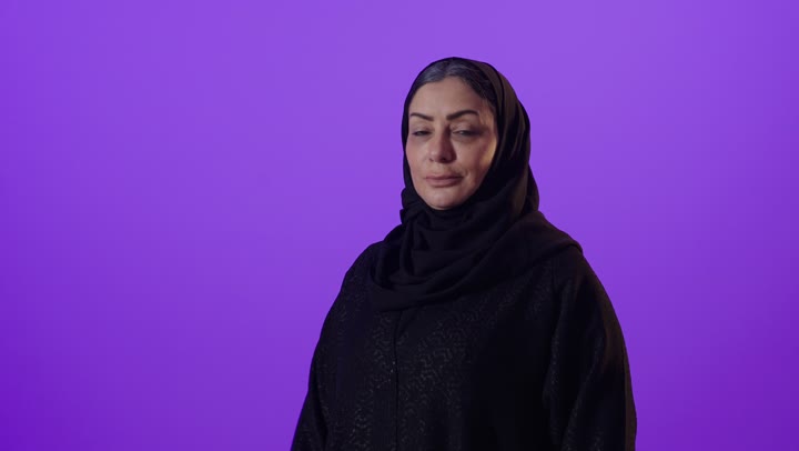 مفهوم الأنوثة والملامح الناعمة، ايماءات الثقة بالنفس، الاناقة والاهتمام بالمظهر الخارجي,  بورتريه لإمرأة كبيرة بالسن عربية خليجية سعودية محجبة ترتدي العباءة تقف بايدي متقاطعة تنظر الى الكاميرا بتعابير السعادة, خلفية بنفسجية