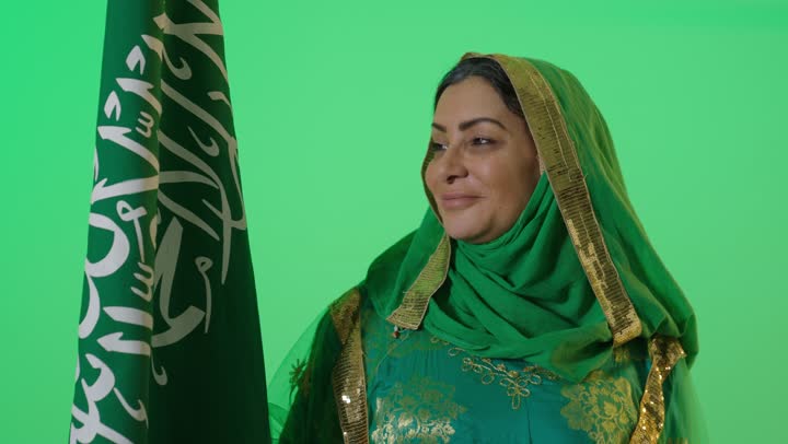 يوم بدينا 1727 م، ذكرى تأسيس الدولة السعودية الأولى 22 فبراير، ارتداء زي المنطقة الوسطى في المناسبات الشعبية والوطنية,  الاعتزاز بإنجازات الوطن، شعار الهوية الوطنية وراية الإسلام والتوحيد، بورتريه لامراه عربيه خليجيه سعوديه كبيرة بالسن ترتدي البرقع والشيلة تحمل بيدها علم التأسيس, تنظر بايماءات الفخر، كروما، خلفية خضراء