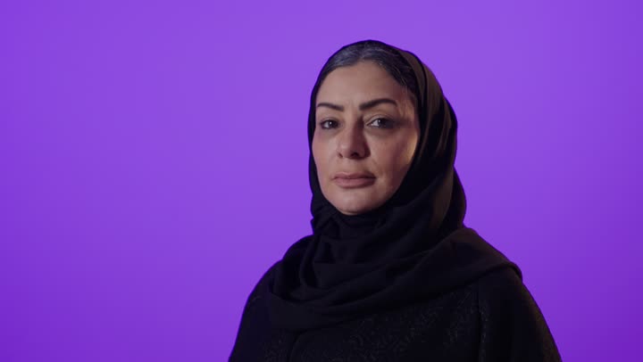 ايماءات الثقة بالنفس، مفهوم الأنوثة والملامح الناعمة، الاناقة والاهتمام بالمظهر الخارجي,  بورتريه لإمرأة كبيرة بالسن عربية خليجية سعودية محجبة ترتدي العباءة تقف بايدي متقاطعة تنظر الى الكاميرا بتعابير السعادة, خلفية بنفسجية