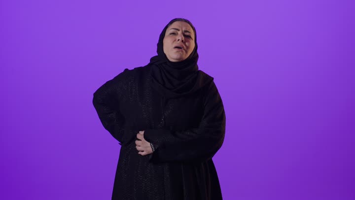 آلام العامود الفقري والظهر، الحاجة إلى العلاج، الشعور بالوجع والألم الجسدي، التعب والإرهاق والإحساس بالمرض،بورتريه لإمرأة كبيرة بالسن عربية خليجية سعودية محجبة ترتدي العباءة تضع يدها على ظهرها ، خلفية بنفسجية