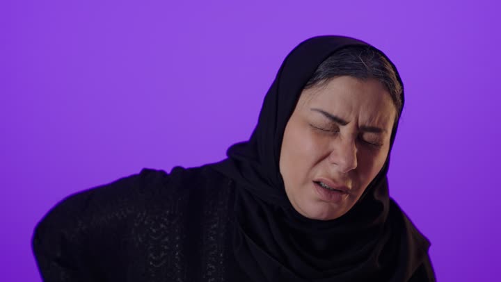 الحاجة إلى العلاج، آلام العامود الفقري والظهر، الشعور بالوجع والألم الجسدي، التعب والإرهاق والإحساس بالمرض،بورتريه لإمرأة كبيرة بالسن عربية خليجية سعودية محجبة ترتدي العباءة تضع يدها على ظهرها ، خلفية بنفسجية
