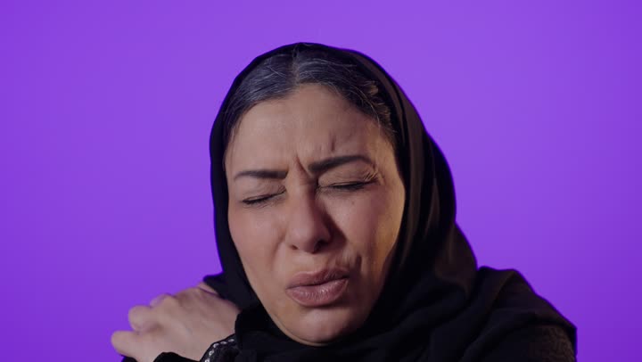 الآلام والأوجاع العصبية والعضلية، التهاب العضلات والمفاصل، الاحساس بالمرض والتعب، بورتريه لإمرأة كبيرة بالسن عربية خليجية سعودية محجبة ترتدي العباءة تضع يدها على كتفها بإيماءات تدل على الألم، خلفية بنفسجية