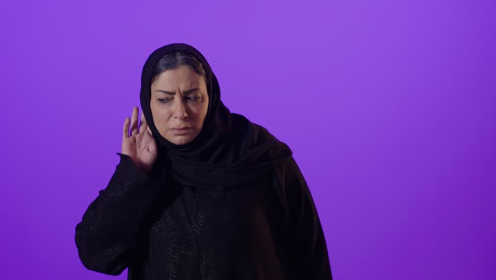 بورتريه لإمرأة كبيرة بالسن عربية خليجية سعودية محجبة ترتدي العباءة تضع يدها خلف أذنها بإيماءات التنصت والتجسس، التركيز على الكلام وسماع الاسرار، الانصات وحسن الاستماع، خلفية بنفسجية