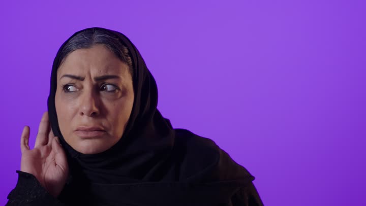 الانصات وحسن الاستماع، بورتريه لإمرأة كبيرة بالسن عربية خليجية سعودية محجبة ترتدي العباءة تضع يدها خلف أذنها بإيماءات التنصت والتجسس، التركيز على الكلام وسماع الاسرار، خلفية بنفسجية