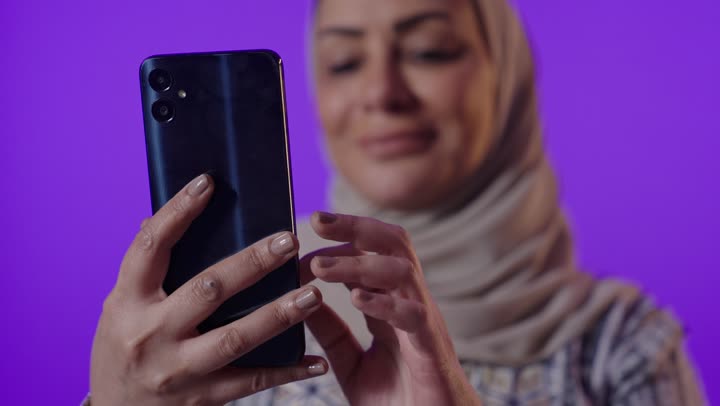 Browsing applications and social media programs, exchanging conversations and news with family and friends, a portrait of an elderly Arab Gulf Saudi woman wearing a hijab and an abaya holding a mobile phone, using modern advanced devices and technologies, with a purple background.