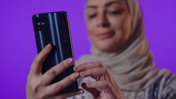 تصفح التطبيقات وبرامج التواصل الاجتماعي، تبادل الأحاديث والأخبار مع الأهل والأصدقاء، بورتريه لإمرأة كبيرة بالسن عربية خليجية سعودية محجبة ترتدي العباءة تمسك بيدها الهاتف المحمول، استخدام الأجهزة والتقنيات الحديثة المتطورة، خلفية بنفسجية