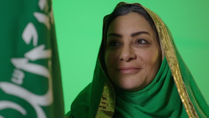 يوم بدينا 1727 م، ذكرى تأسيس الدولة السعودية الأولى 22 فبراير، ارتداء زي المنطقة الوسطى في المناسبات الشعبية والوطنية,  الاعتزاز بإنجازات الوطن، شعار الهوية الوطنية وراية الإسلام والتوحيد، بورتريه لامراه عربيه خليجيه سعوديه كبيرة بالسن ترتدي البرقع والشيلة تحمل بيدها علم التأسيس, تنظر بايماءات الفخر، كروما، خلفية خضراء