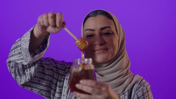 غذاء طبيعي صحي، منتج  وطني محلي سعودي،النظر إلى الكاميرا بايماءات السرور والسعادة، بورتريه لإمرأة كبيرة بالسن عربية خليجية سعودية محجبة ترتدي العباءة تحمل بيدها عبوة زجاجية من عسل النحل الطازج، خلفية بنفسجية