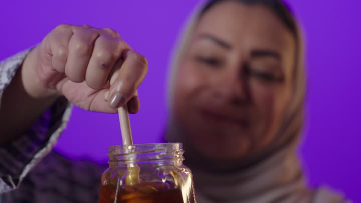 منتج  وطني محلي سعودي، غذاء طبيعي صحي، النظر إلى الكاميرا بايماءات السرور والسعادة، بورتريه لإمرأة كبيرة بالسن عربية خليجية سعودية محجبة ترتدي العباءة تحمل بيدها عبوة زجاجية من عسل النحل الطازج، خلفية بنفسجية