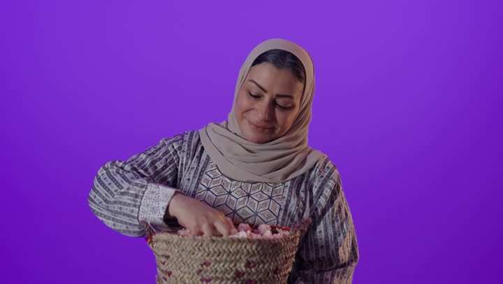 الزهور والورود العطرية، النظر  بايماءات السرور والسعادة، بورتريه لإمرأة كبيرة بالسن عربية خليجية سعودية محجبة ترتدي العباءة تحمل بيدها سلة قش ممتلئة بالورد الجوري الطبيعي، خلفية بنفسجية