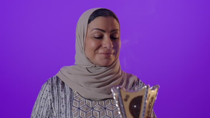 التطيب والتعطر بالبخور, أصالة العادات والتقاليد الخليجية, استخدام خشب العود بالأعياد والمناسبات, بورتريه لإمرأة كبيرة بالسن عربية خليجية سعودية محجبة ترتدي العباءة  تمسك المبخرة التراثية المزخرفة بيدها ، تنظر بإيماءات السعادة و السرور, خلفية بنفسجية