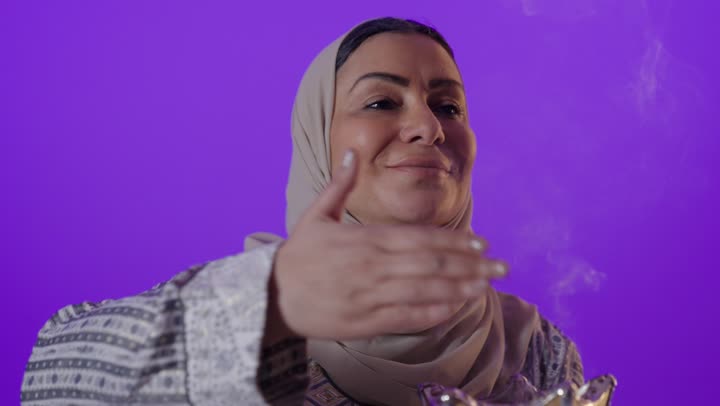 بورتريه لإمرأة كبيرة بالسن عربية خليجية سعودية محجبة ترتدي العباءة  تمسك المبخرة التراثية المزخرفة بيدها ، تنظر بإيماءات السعادة و السرور, أصالة العادات والتقاليد الخليجية, استخدام خشب العود بالأعياد والمناسبات, التطيب والتعطر بالبخور, خلفية بنفسجية