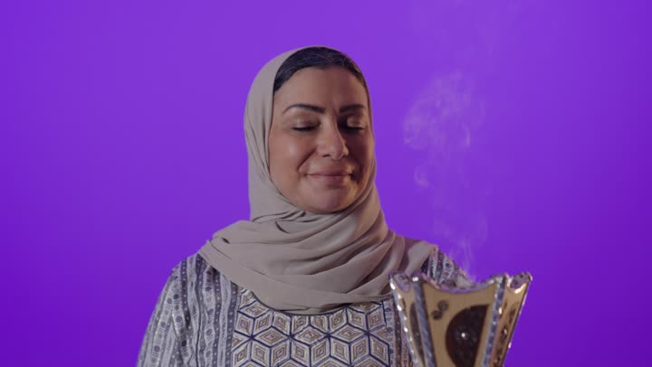 أصالة العادات والتقاليد الخليجية, استخدام خشب العود بالأعياد والمناسبات, بورتريه لإمرأة كبيرة بالسن عربية خليجية سعودية محجبة ترتدي العباءة  تمسك المبخرة التراثية المزخرفة بيدها ، تنظر بإيماءات السعادة و السرور, التطيب والتعطر بالبخور, خلفية بنفسجية