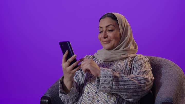 تبادل الأحاديث والأخبار مع الأهل والأصدقاء، تصفح التطبيقات وبرامج التواصل الاجتماعي، بورتريه لإمرأة كبيرة بالسن عربية خليجية سعودية محجبة ترتدي العباءة تمسك بيدها الهاتف المحمول، استخدام الأجهزة والتقنيات الحديثة المتطورة، خلفية بنفسجية
