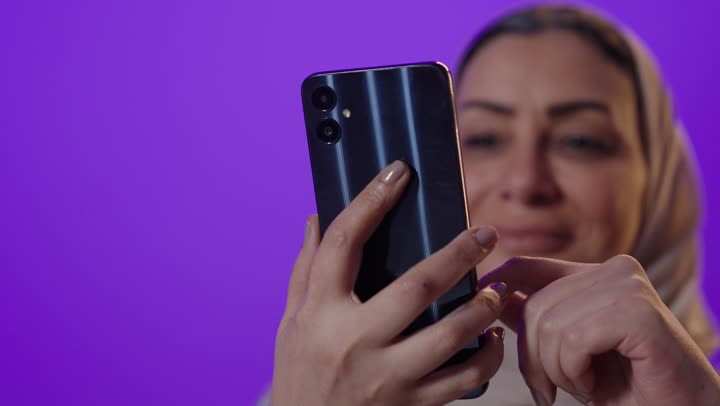Browsing applications and social media programs, exchanging conversations and news with family and friends, a portrait of an elderly Arab Gulf Saudi woman wearing a hijab and an abaya holding a mobile phone, using modern advanced devices and technologies, with a purple background.