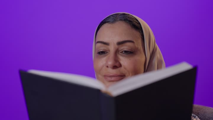 الاسترخاء والاستمتاع بالأجواء الهادئة، الشعور بالراحة والطمأنينة, قراءة الروايات والقصص الممتعة،  بورتريه لإمرأة كبيرة بالسن عربية خليجية سعودية محجبة ترتدي العباءة تمسك بيدها الكتاب، اتباع أسلوب حياة صحي، خلفية بنفسجية