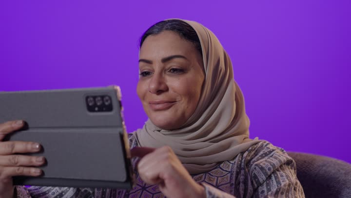 دمج التقنية والتكنولوجيا الحديثة في الحياة اليومية، مشاهدة الافلام والمسلسلات ، ايماءات الاندماج والتركيز لإنجاز المهام ، ادارة العمل عن بعد ، إستخدام الجهاز اللوحي ، بورتريه لإمرأة كبيرة بالسن عربية خليجية سعودية محجبة ترتدي العباءة تستخدم التابلت ،  تصفح الخدمات والتطبيقات عبر الانترنت ، خلفية بنفسجية.