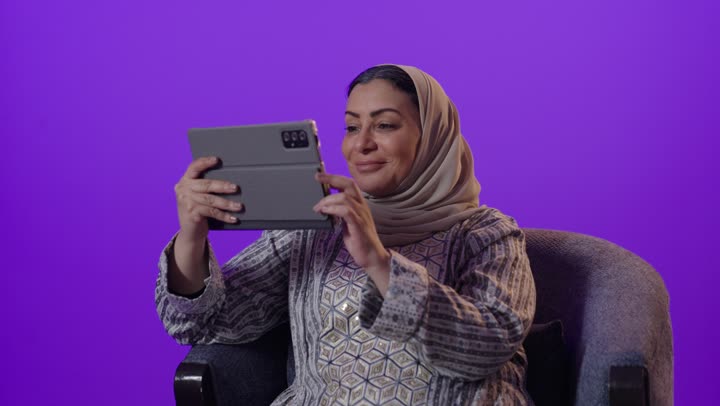 Watching movies and series, integrating modern technology and technology into daily life, gestures of integration and focus to accomplish tasks, managing remote work, using a tablet, a portrait of an elderly Arab Gulf Saudi woman wearing a hijab and an abaya using a tablet, browsing services and applications online, purple background.