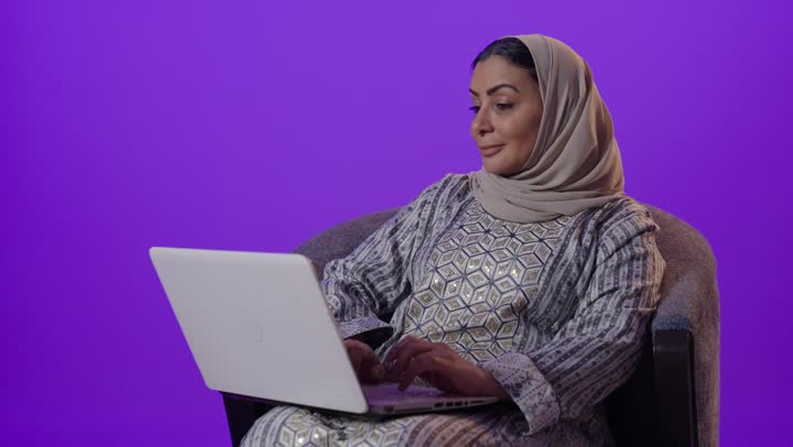 Remote work management, integrating modern technology and technology into daily life, gestures of engagement and focus to accomplish tasks, using a laptop, a portrait of an elderly Arab Gulf Saudi woman wearing a hijab and an abaya using a laptop, browsing services and applications online, the concept of digital advertising and marketing, purple background.