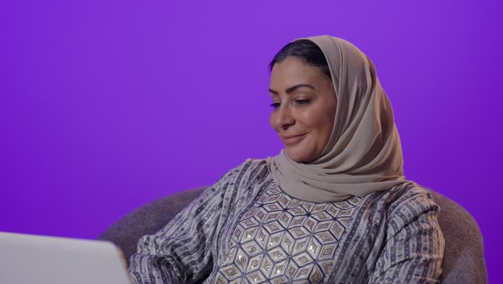Gestures of integration and focus to accomplish tasks, integrating modern technology and technology into daily life, managing remote work, using a laptop, a portrait of an elderly Arab Gulf Saudi woman wearing a hijab and an abaya using a laptop, browsing services and applications online, the concept of digital advertising and electronic marketing, purple background.