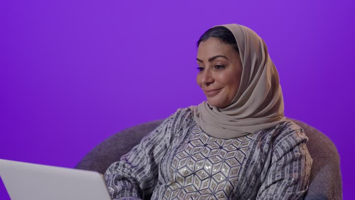 Integrating modern technology and technology into daily life, gestures of integration and focus to accomplish tasks, managing remote work, using a laptop, a portrait of an elderly Arab Gulf Saudi woman wearing a hijab and an abaya using a laptop, browsing services and applications online, the concept of digital advertising and electronic marketing, purple background.