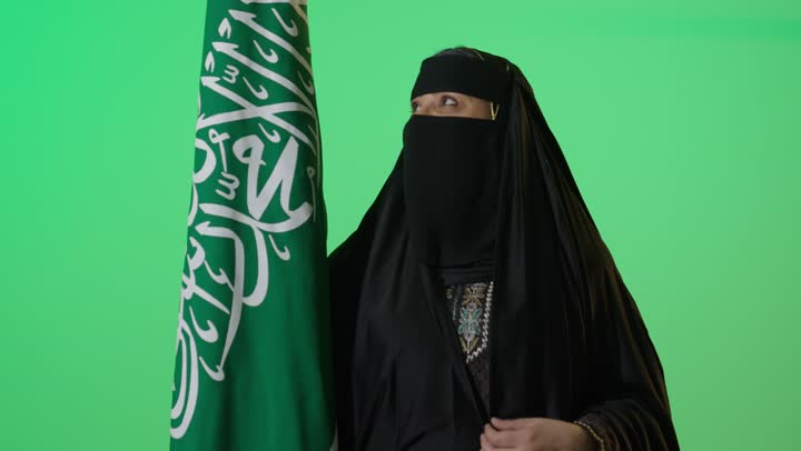 ذكرى تأسيس الدولة السعودية الأولى 22 فبراير، شعار الهوية الوطنية وراية الإسلام والتوحيد، بورتريه لامراه عربيه خليجيه سعوديه منتقبة كبيرة بالسن ترتدي العباءة السوداء تحمل بيدها علم التأسيس, تنظر بايماءات الفخر،  الاعتزاز بإنجازات الوطن، كروما، خلفية خضراء