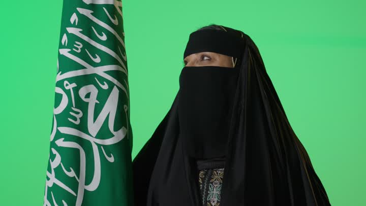 شعار الهوية الوطنية وراية الإسلام والتوحيد، بورتريه لامراه عربيه خليجيه سعوديه منتقبة كبيرة بالسن ترتدي العباءة السوداء تحمل بيدها علم التأسيس, تنظر بايماءات الفخر، ذكرى تأسيس الدولة السعودية الأولى 22 فبراير، الاعتزاز بإنجازات الوطن، كروما، خلفية خضراء