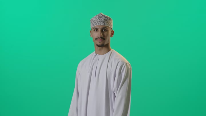 بورتريه لرجل عربي عماني شايب يرتدي الدشداشة والكمه يقف بشكل متكتف ينظر إلى الكاميرا بتعابير الابتسامة، وضع اليدين على الذراعين وشد الظهر, الوقوف باستقامة مع تقاطع الايدي، خلفية خضراء