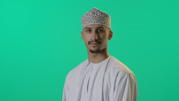 الوقوف باستقامة مع تقاطع الايدي، بورتريه لرجل عربي عماني شايب يرتدي الدشداشة والكمه يقف بشكل متكتف ينظر إلى الكاميرا بتعابير الابتسامة، وضع اليدين على الذراعين وشد الظهر خلفية خضراء