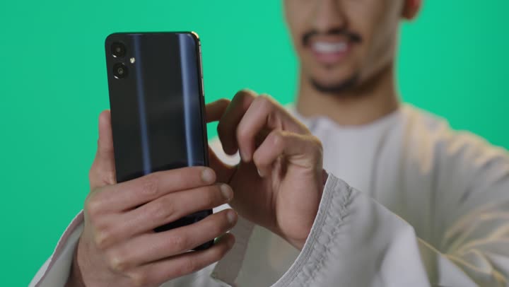 بورتريه مقرب ليد رجل عربي خليجي عماني يرتدي الدشداشة والكمة يستخدم الهاتف المحمول، الدردشة مع الأهل والأصدقاء، تصفح مواقع التواصل الاجتماعي، استخدام جهاز تقني حديث, دمج التكنولوجيا المتطورة بالحياة اليومية، خلفية خضراء