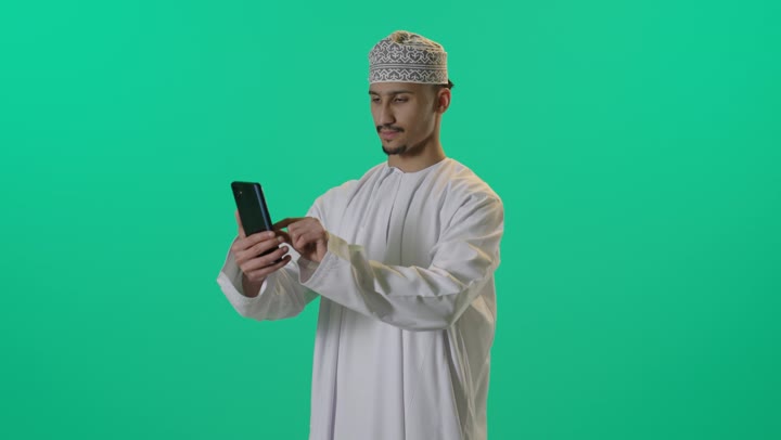Chatting with family and friends, browsing social media, using modern technology, integrating advanced technology into daily life, a portrait of an Omani Gulf Arab man wearing a dishdasha and kumma using a mobile phone, green background.