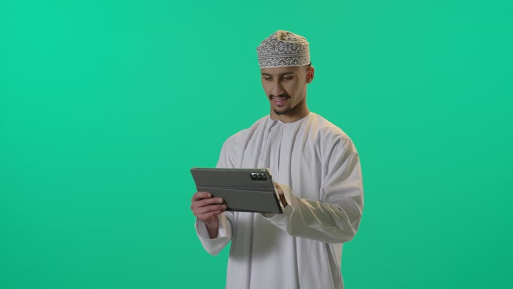 The use of advanced technology, a close-up portrait of a young Arab Gulf Omani man wearing a dishdasha and kumma, holding a tablet device in his hand, chatting and communicating with family and friends, browsing social media, with a green background.