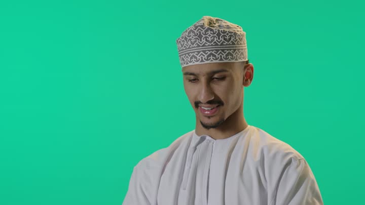The use of advanced technology, a close-up portrait of a young Arab Gulf Omani man wearing a dishdasha and kumma, holding a tablet device in his hand, chatting and communicating with family and friends, browsing social media, with a green background.