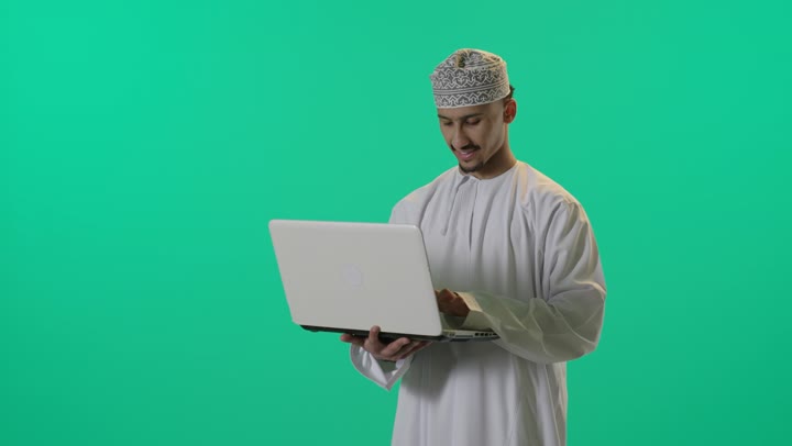 انجاز مهام العمل، تصفح الانترنت، احتراف وظائف الحاسوب وتكنولوجيا المعلومات، دردشة رقمية، استخدام وسائل تقنية حديثة, بورتريه لرجل عربي خليجي عماني يحمل بيده جهاز الحاسوب المحمول، العمل عن بعد، خلفية خضراء