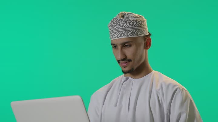 بورتريه لرجل عربي خليجي عماني يحمل بيده جهاز الحاسوب المحمول، العمل عن بعد، انجاز مهام العمل، تصفح الانترنت، احتراف وظائف الحاسوب وتكنولوجيا المعلومات، دردشة رقمية، استخدام وسائل تقنية حديثة, خلفية خضراء