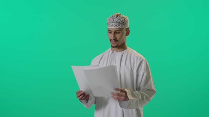 بورتريه لرجل عربي خليجي عماني يرتدي الدشداشة والكمة يحمل بيده كراسه تدوين الملاحظات يقوم بتصفح الاوراق، اعداد تقارير ومهام العمل، دراسة جدوى الاعمال, مراجعة نتائج الاجتماع، خلفية خضراء